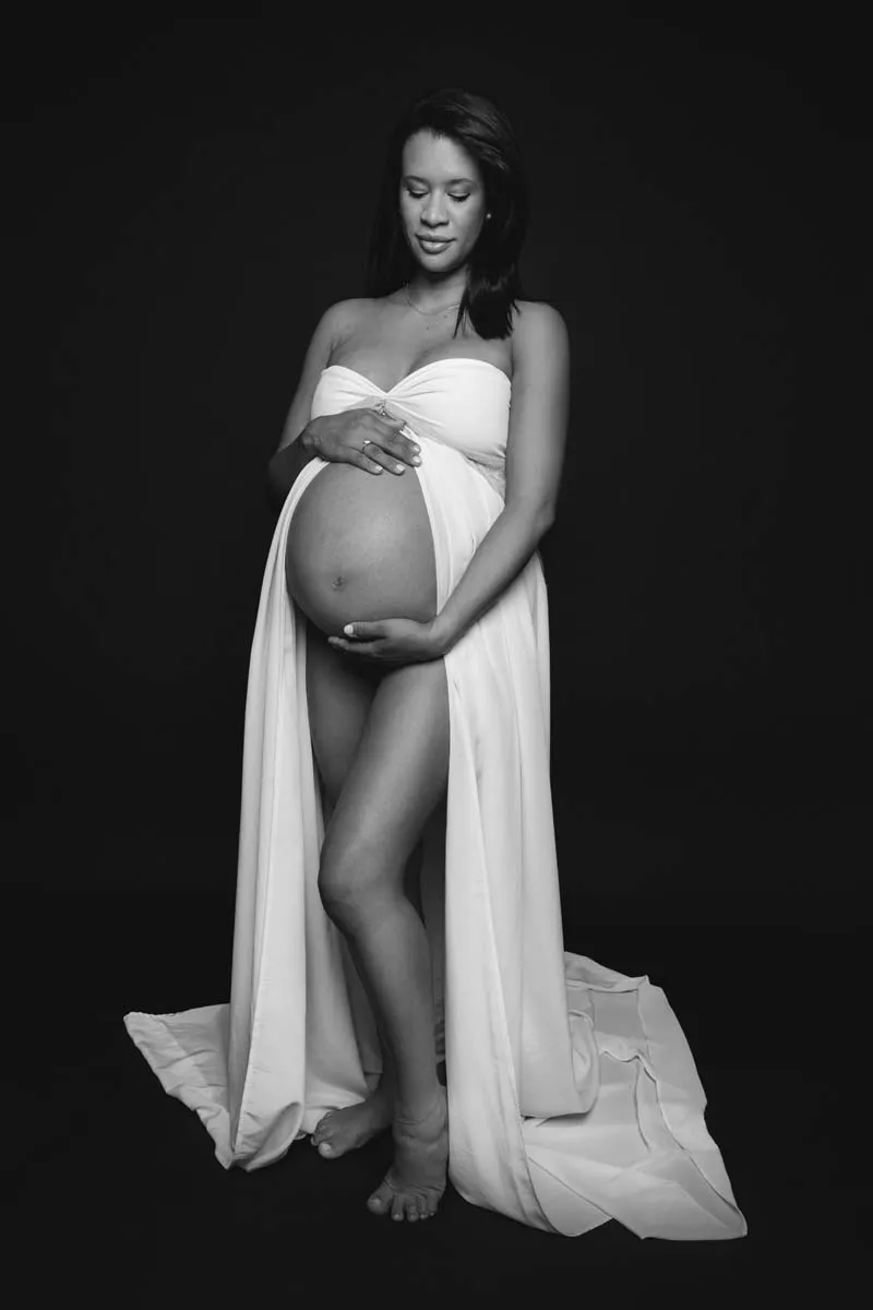 Sesión premamá clásica con vestido blanco sobre fondo oscuro, en blanco y negro