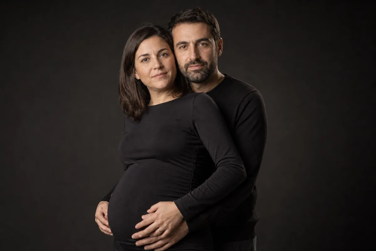 Pareja premamá en el estudio, él abrazándola por detrás con las manos unidas